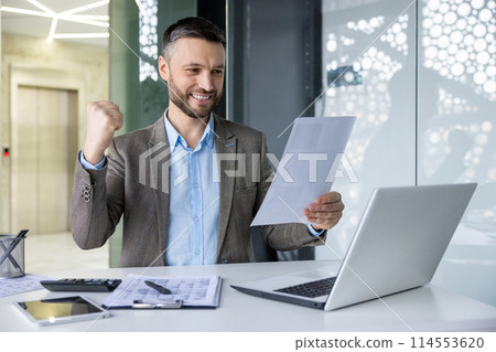 Cheerful businessman celebrating achievement in modern office environment, looking at documents while working on laptop Cheerful businessman celebrating achievement in modern office environment, looking at documents while working on laptop 114553620