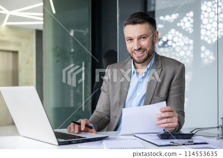 Smiling businessman at his desk working on laptop and checking documents in a contemporary office setting. 114553635