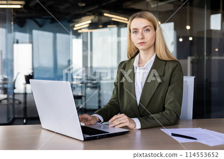 Confident businesswoman wearing a green blazer, focusing on her laptop at a modern office desk, surrounded by glass walls. 114553652