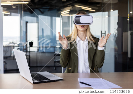 A professional young businesswoman experiences virtual reality with a VR headset in a sleek, modern office setting, surrounded by high-tech ambiance. 114553656