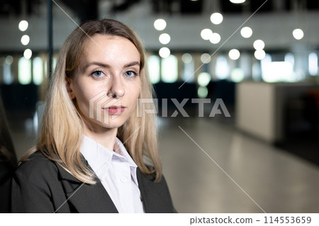 A professional portrait of a young blonde businesswoman standing confidently in a modern office environment, exuding leadership and sophistication. 114553659