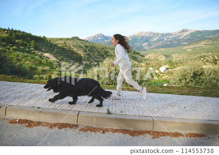 Full length side portrait of a cute child girl running while walking her dog on leash on the nature outdoors Full length side portrait of a cute child girl running while walking her dog on leash on the nature outdoors 114553738