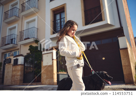 Caucasian adorable little girl taking her dog for a walk on leash on the city street on a sunny day, dressed in white sportswear. Lovely kid walking her cocker spaniel outdoors Caucasian adorable little girl taking her dog for a walk on leash on the city street on a sunny day, dressed in white sportswear. Lovely kid walking her cocker spaniel outdoors 114553751