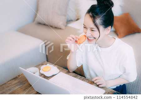 Young woman eating a snack while looking at a computer Young woman eating a snack while looking at a computer 114554842