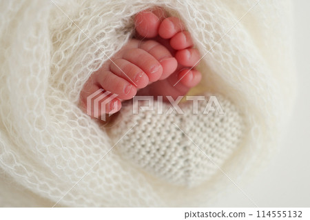 The tiny foot of a newborn baby. Soft feet of a new born in a white wool blanket. Close up of toes, heels and feet of a newborn. Knitted white heart in the legs of a baby. Macro photography The tiny foot of a newborn baby. Soft feet of a new born in a white wool blanket. Close up of toes, heels and feet of a newborn. Knitted white heart in the legs of a baby. Macro photography 114555132