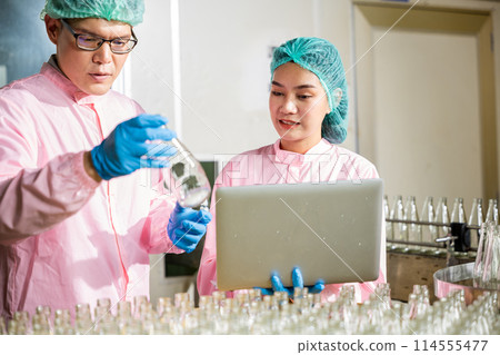 Within a beverage factory line engineers meticulously inspect product bottles on the conveyor belt. Inspection and quality control managed by professionals using a laptop. 114555477