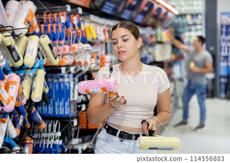 Female buyer selects necessary items for repair and carefully examines painting roller for renovations. Female buyer selects necessary items for repair and carefully examines painting roller for renovations. 114556883