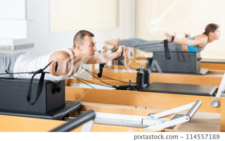 Concentrated man performing exercises with straps on Pilates reformer 114557189
