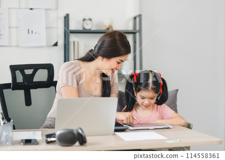 Young mother working on laptop beside her daughter playing on tablet at home office. 114558361