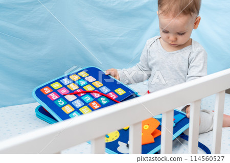 Little baby girl playing with busy book while sitting in crib. Concept of quiet books and modern educational toys Little baby girl playing with busy book while sitting in crib. Concept of quiet books and modern educational toys 114560527