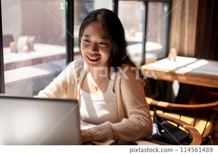 Young positive Asian woman working remotely at a coffee shop, working on her laptop computer. Young positive Asian woman working remotely at a coffee shop, working on her laptop computer. 114561489