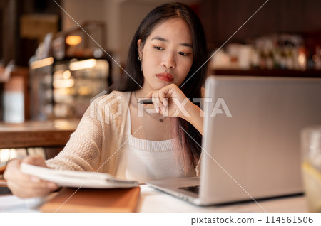 Young focused Asian woman working remotely at a coffee shop, taking notes in her notebook. 114561506