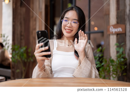 A cheerful Asian woman enjoys talking on a video call while sitting at an outdoor table of a cafe. A cheerful Asian woman enjoys talking on a video call while sitting at an outdoor table of a cafe. 114561528