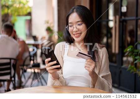 A happy Asian woman sits at an outdoor table of a cafe, holding her credit card and smartphone. A happy Asian woman sits at an outdoor table of a cafe, holding her credit card and smartphone. 114561532