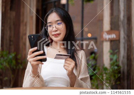 A happy Asian woman sits at an outdoor table of a cafe, holding her credit card and smartphone. 114561535