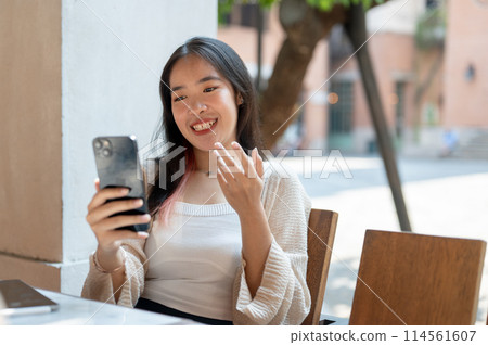 A cheerful Asian woman enjoys talking on a video call while chilling at a restaurant in the city. 114561607