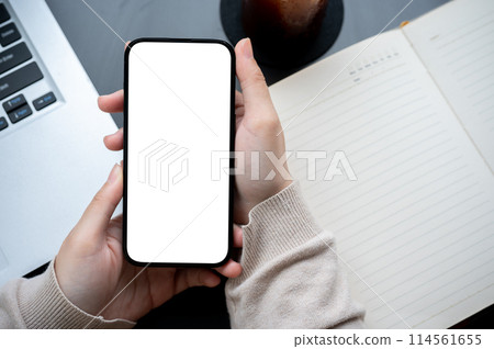 Top view image of a woman's hand holding a white-screen smartphone mockup over a modern office desk. 114561655