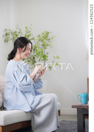 A young woman thinking with her smartphone in a beautiful living room, looking at the screen and finding something with a smile, vertical 114562523