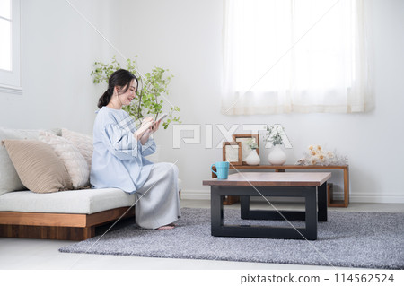 一位年輕女子在美麗的客廳裡拿著智慧型手機思考,廣角全身看著螢幕,微笑著找到一些東西。 一位年輕女子在美麗的客廳裡拿著智慧型手機思考,廣角全身看著螢幕,微笑著找到一些東西。 114562524