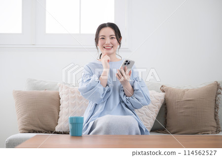 A young woman thinking with her smartphone in a beautiful living room, looking at the screen and finding something with a smile, front view, looking at the camera 114562527