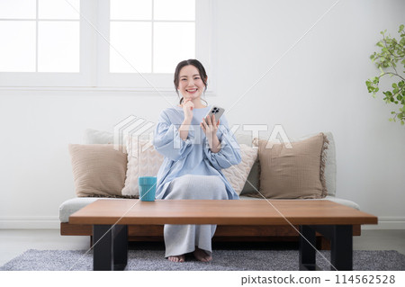 A young woman thinking with her smartphone in a beautiful living room, looking at the screen and finding something with a smile, wide angle, front, looking at the camera 114562528