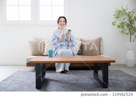 A young woman thinking with her smartphone in a beautiful living room, looking at the screen and finding something with a smile, wide angle, front, looking at the camera 114562529