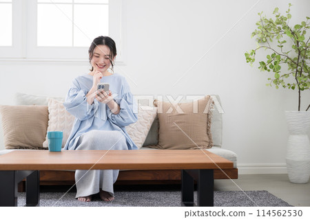 A young woman thinking with her smartphone in a beautiful living room, looking at the screen and finding something with a smile, wide-angle full body, front A young woman thinking with her smartphone in a beautiful living room, looking at the screen and finding something with a smile, wide-angle full body, front 114562530