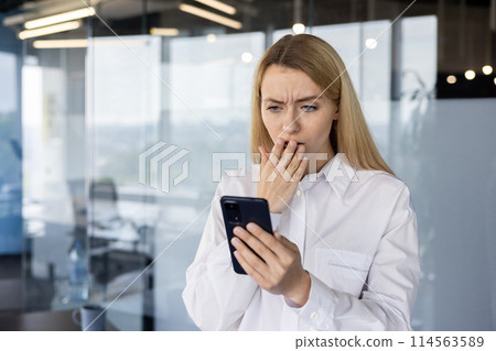 A young businesswoman stands in a modern office, shocked and concerned as she reads a surprising text message on her smartphone. A young businesswoman stands in a modern office, shocked and concerned as she reads a surprising text message on her smartphone. 114563589