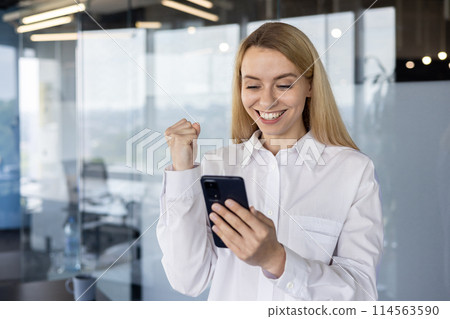 A joyful businesswoman in a modern office successfully completes a task and celebrates smiling while looking at her smartphone. A joyful businesswoman in a modern office successfully completes a task and celebrates smiling while looking at her smartphone. 114563590