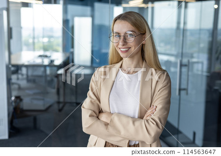 A professional, confident businesswoman stands with arms crossed in a bright, modern office setting, showcasing a smart and approachable demeanor. 114563647