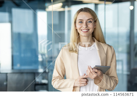 A professional businesswoman with a pleasant smile uses a digital tablet in a contemporary office environment, exuding confidence and competence. 114563671