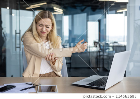A young professional woman experiences discomfort in her arm during laptop use in a modern office setting, indicating potential repetitive strain injury. 114563695