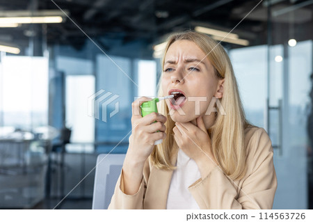 A woman in an office uses a throat spray for sore throat relief. She appears to be in discomfort while medicating in a modern workspace. A woman in an office uses a throat spray for sore throat relief. She appears to be in discomfort while medicating in a modern workspace. 114563726