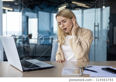 A professional businesswoman experiencing severe ear pain, possibly otitis, while working in a contemporary office setting. A professional businesswoman experiencing severe ear pain, possibly otitis, while working in a contemporary office setting. 114563743