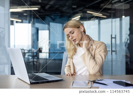 Young businesswoman feeling neck pain while working on laptop at office desk. Concept of work-related stress and discomfort. Young businesswoman feeling neck pain while working on laptop at office desk. Concept of work-related stress and discomfort. 114563745