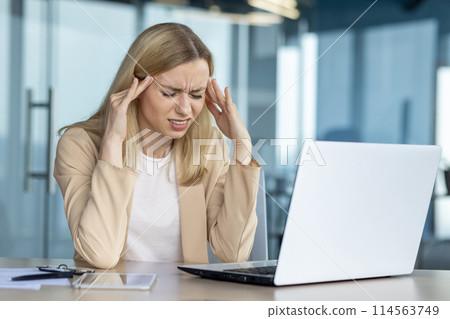 A young businesswoman experiences a painful headache while working at her laptop in a modern office. She appears stressed and overwhelmed. 114563749