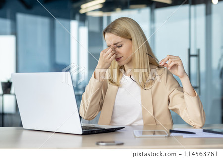 Businesswoman feeling stressed and fatigued while working on her laptop in a modern office setting. She holds her glasses and rubs her eyes due to exhaustion. Businesswoman feeling stressed and fatigued while working on her laptop in a modern office setting. She holds her glasses and rubs her eyes due to exhaustion. 114563761