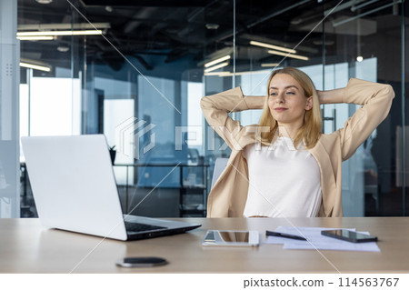 A professional woman takes a break at her workplace, stretching with contentment. A modern office space serves as the perfect backdrop for productivity and relaxation. 114563767
