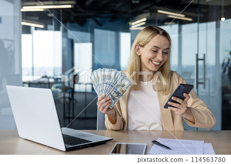 A happy woman in an office holding a stack of cash and smiling at her phone. She sits at a desk with a laptop, portraying success and financial achievement. A happy woman in an office holding a stack of cash and smiling at her phone. She sits at a desk with a laptop, portraying success and financial achievement. 114563780