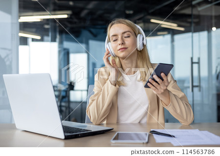 Businesswoman enjoying music on headphones and smartphone while working on a laptop in a modern office setting. 114563786