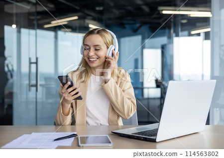Happy young businesswoman listens to music on headphones while using smartphone and laptop in a bright office setting. Enjoying a small break during work. 114563802