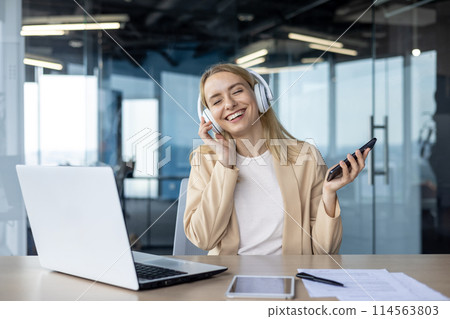 Happy businesswoman enjoying music with headphones while working at the office. She is smiling and holding a smartphone. Laptop and tablet on the desk. 114563803