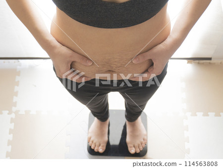 A woman weighing herself while pinching the fat on her belly, which still shows stretch marks after giving birth 114563808