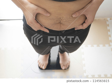A woman weighing herself while pinching the fat on her belly, which still shows stretch marks after giving birth 114563815