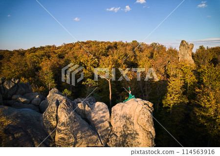 Woman practicing yoga and meditation on the top of huge boulder in the evening. Beautiful sunset, autumn forests, rocks and hills on background. Aerial drone view Woman practicing yoga and meditation on the top of huge boulder in the evening. Beautiful sunset, autumn forests, rocks and hills on background. Aerial drone view 114563916