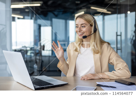 A cheerful professional woman engaging in a video call while working on her laptop in a brightly lit, contemporary office space. A cheerful professional woman engaging in a video call while working on her laptop in a brightly lit, contemporary office space. 114564077