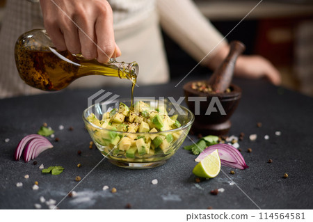 Woman pours olive oil onto Chopped ingredients in wooden bowl - avocado, onion and pepper 114564581
