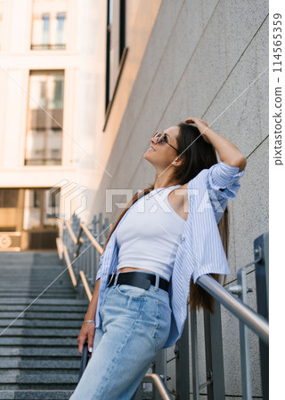 Happy woman in a white tank top and blue jeans is leaning against a railing. She is smiling and she is enjoying herself Happy woman in a white tank top and blue jeans is leaning against a railing. She is smiling and she is enjoying herself 114565359