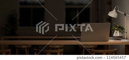 A dark classroom or co-working space at night features a laptop computer on a wooden table. A dark classroom or co-working space at night features a laptop computer on a wooden table. 114565457