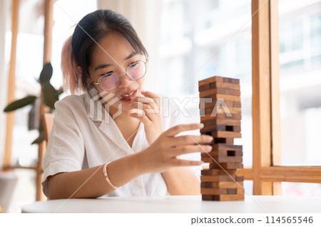 A young Asian woman focusing on playing a wooden block game at a table indoors. 114565546